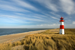 Der nördlichste Leuchtturm Deutschlands auf dem Sylter Ellenbogen, umgeben von Dünen und der Nordsee, unter einem weiten blauen Himmel.