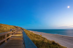 Holzsteg führt durch Dünenlandschaft hinunter zum Strand bei Abenddämmerung.