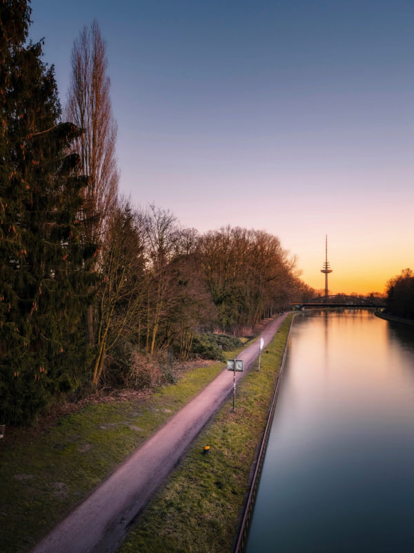 Blick auf den friedvollen Dortmund-Ems-Kanal in Münster bei Sonnenuntergang mit dem Fernsehturm im Hintergrund und einem idyllischen Wanderweg am Ufer.