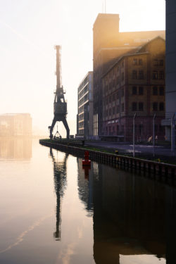 Morgendliches Gegenlicht umreißt den Hafen-Kran und Gebäude am Stadthafen Münster, mit Reflexionen im Wasser.