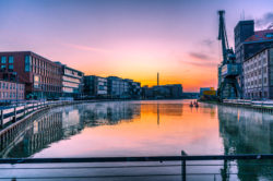 Panoramablick auf den Stadthafen Münster mit modernen Gebäuden, spiegelndem Wasser bei Sonnenaufgang und einem markanten Hafen-Kran im Vordergrund.