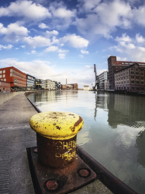 Ein verwitterter, gelber Poller im Vordergrund mit Blick auf den Stadthafen von Münster und den Hafen-Kran, fotografiert von Thomas Branse.