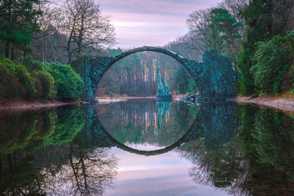 Steinbogen-Brücke, die sich über einen ruhigen See erstreckt, umgeben von Bäumen und reflektiert im Wasser.