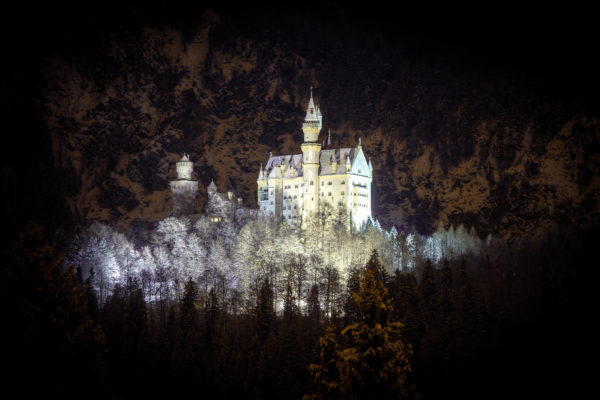 Beleuchtetes Schloss Neuschwanstein im Winter vor dunklem Bergpanorama und verschneiten Bäumen.