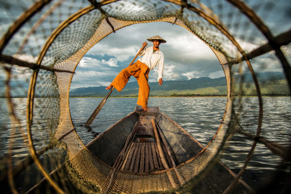 Fischer in traditioneller Kleidung balanciert auf einem Boot, gesehen durch ein Fischernetz, mit majestätischen Bergen im Hintergrund.