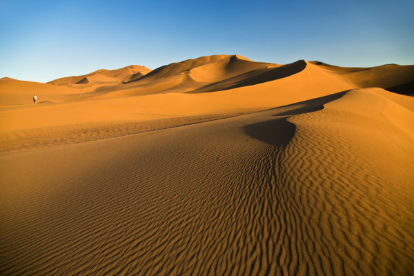 Ein einsamer Wanderer überquert eine ausgedehnte goldene Wüstendüne bei Sonnenuntergang.