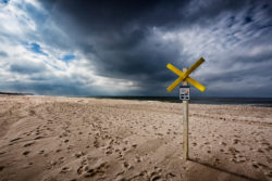 Warnschild vor gefährlichen Buhnenresten am Sylter Strand mit dramatischem Wolkenhimmel im Hintergrund.