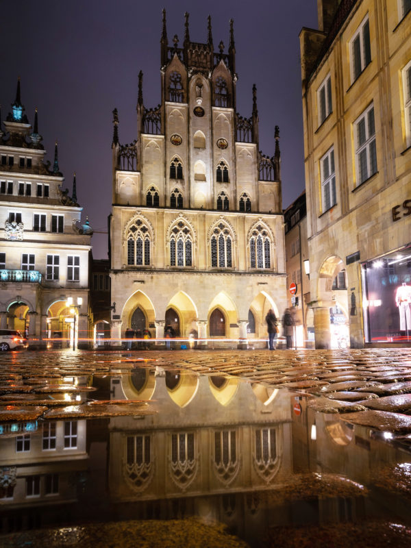 Nachtansicht des Historischen Rathauses von Münster mit Spiegelung auf nasser Oberfläche, belebte Altstadtatmosphäre im Prinzipalmarkt.