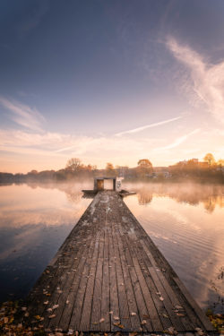 Sonnenaufgang über dem Aasee in Münster mit einem Holzsteg, der ins Wasser führt, umgeben von Herbstlaub und einem urbanen Hintergrund.