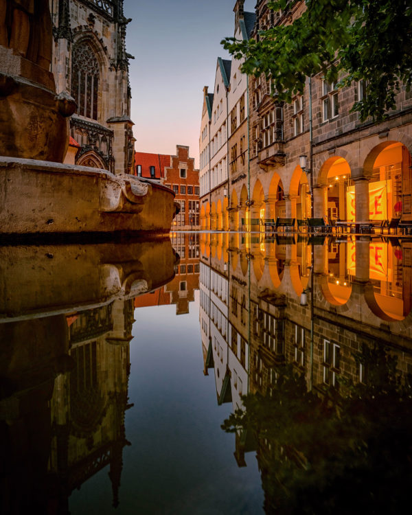 Abendliche Spiegelung des Lambertus-Brunnens und der St. Lamberti-Kirche auf dem Lamberti-Kirchplatz in Münster, beleuchtete Altstadtfassaden spiegeln sich im Wasser.
