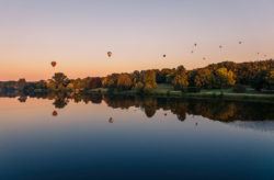 Aasee in Münster bei Sonnenuntergang, spiegelnde Wasseroberfläche, farbenfroher Himmel, Montgolfiade-Ballons im Hintergrund Beschriftung: Münster Aasee Sonnenuntergang - Montgolfiad