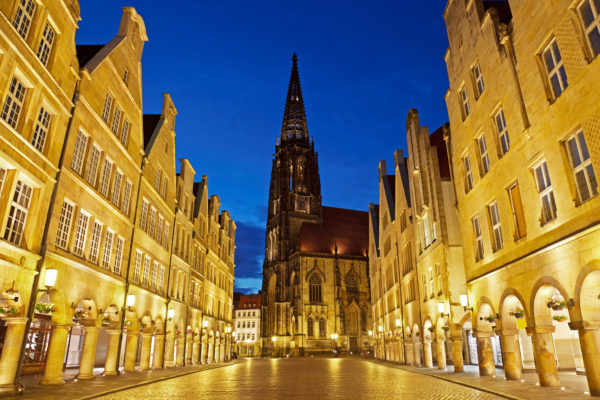 Nächtliche Ansicht des Prinzipalmarkts in Münster mit beleuchteten historischen Gebäuden und dem Kirchturm von St. Lamberti im Hintergrund.