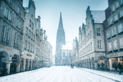 Der Prinzipalmarkt in Münster bedeckt mit Schnee im Winter, beleuchtet von Straßenlaternen mit der Silhouette von St. Lamberti im Hintergrund.