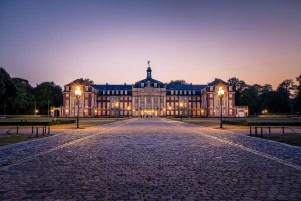 Abendansicht des Fürstbischöflichen Schlosses Münster mit beleuchteter Fassade, eingerahmt von Bäumen und einem gepflasterten Vorplatz.