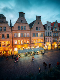 Foto des Prinzipalmarkts in Münster bei Abenddämmerung mit Kopsteinpflaster im Vordergrund, beleuchteten Arkaden im Mittelgrund und abendlichem Himmel im Hintergrund.