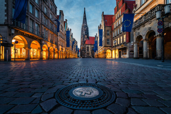 Kontrastreiches Foto des Prinzipalmarkts in Münster mit der Lambertikirche im Hintergrund, umgeben von historischen Gebäuden mit Europafahnen.